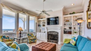 A spacious living room with a large window, a fireplace, a television, and a ceiling fan. - You will delight in the fabulous gulf views from this elegant, Nantucket style beach home! (St. George Island)
