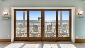 A room with a large wooden door leading to a deck with white chairs and a view of the ocean.