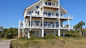 A multi-story beachfront house with balconies, a wooden deck, and a clear view of the ocean.