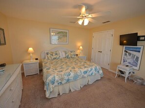 Une chambre à coucher avec un lit, deux tables de chevet, un ventilateur de plafond, une porte et un téléviseur fixé au mur.