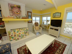 Een woonkamer met een witte open haard, een televisie en uitzicht op het strand door grote ramen.