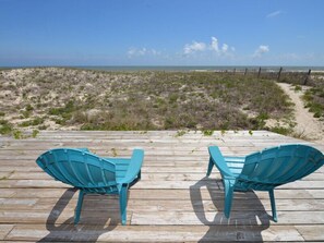 Dos sillas Adirondack turquesa en una terraza de madera con vista a una playa con vegetación escasa y cielo despejado.