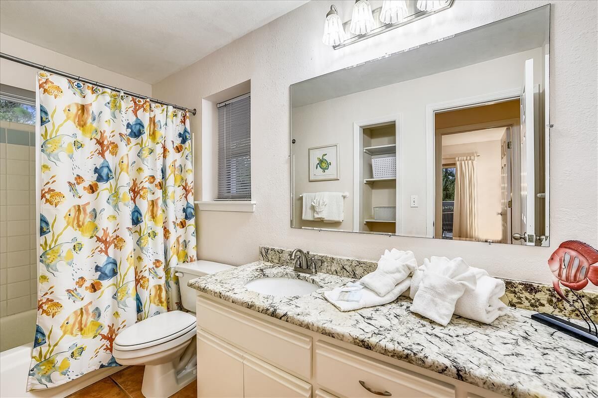 Une salle de bain avec un rideau de douche orné d’un motif de poissons, un lavabo avec un plan de travail en marbre, des toilettes et un miroir au-dessus du lavabo.