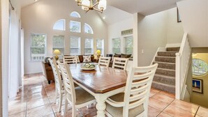 Ruime eetruimte met een grote houten tafel en witte stoelen, een kroonluchter en een trap naar een tweede verdieping.