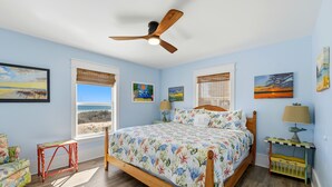 Une chambre à coucher avec un lit en bois, un ventilateur de plafond, une fenêtre avec des stores et des œuvres d’art accrochées aux murs.