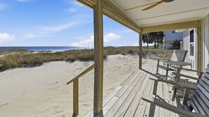Une terrasse en bois dotée d’un ventilateur de plafond, offrant une vue sur une plage avec des dunes de sable et un ciel bleu dégagé.