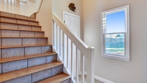 A staircase with wooden steps and white railings, a white door, and a window with a view of a body of water and a distant building. - LEAVE THE city behind and enjoy your escape in this urban designed beach home. (St. George Island)