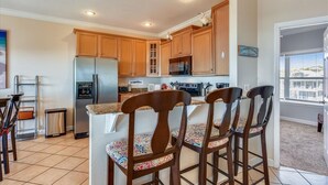 A kitchen with wooden cabinets, a stainless steel refrigerator, a microwave, and a gas stove. There are four wooden chairs with patterned cushions. - LEAVE THE city behind and enjoy your escape in this urban designed beach home. (St. George Island)
