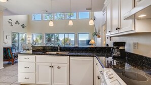 Cocina amplia con una isla grande, armarios blancos y encimera de granito negro. Hay varias lámparas colgantes, una cafetera y vistas al exterior a través de amplias ventanas.