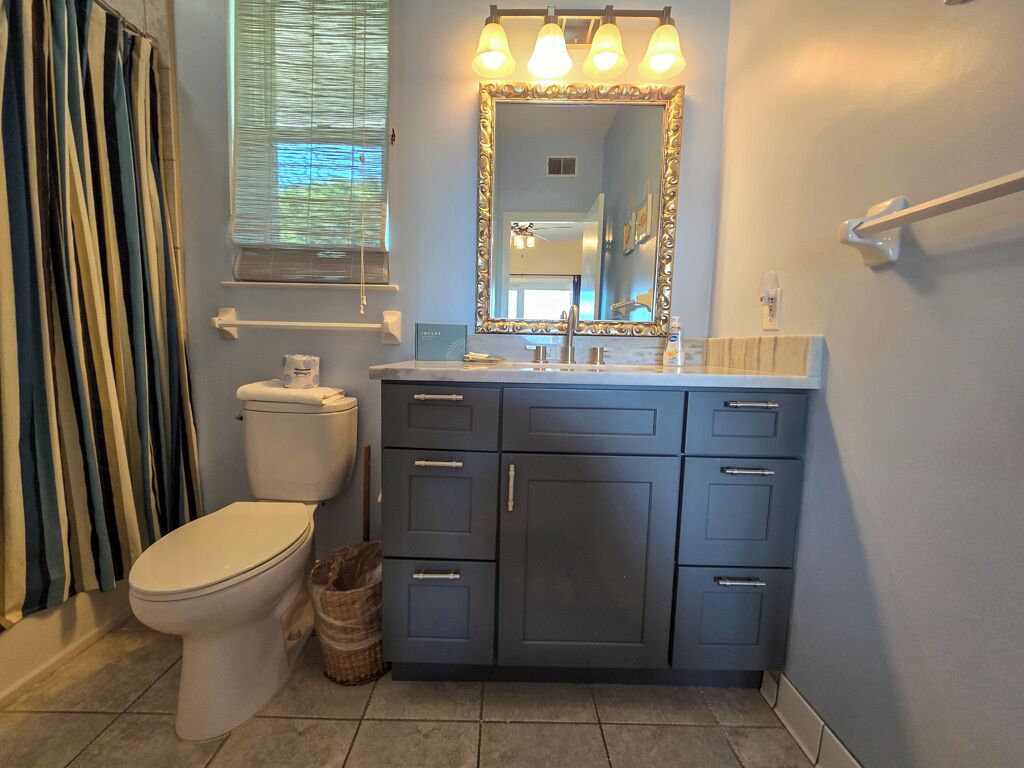 A bathroom with a toilet, a vanity with drawers, a mirror with lights, and a window with blinds.