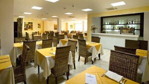 A hotel dining area with multiple tables and wicker chairs, a bar with shelves of bottles, and a white reception desk. - Hotel Club Ravezzo (Ugento)