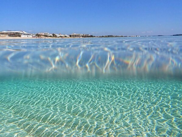 On aperçoit clairement une plage, une mer calme et, au loin, une côte bordée de bâtiments.