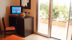 A room with a dark wooden desk, a television, a chair, and a view of a balcony with potted plants. - Hotel Club Ravezzo (Ugento)