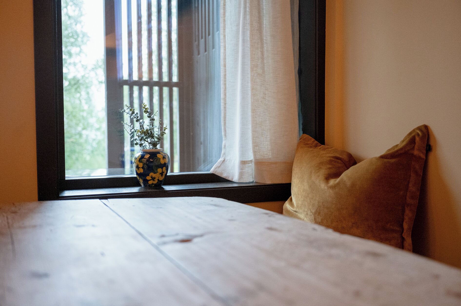 A room with a wooden bed, a window with vertical blinds, a vase with flowers, and a brown pillow.