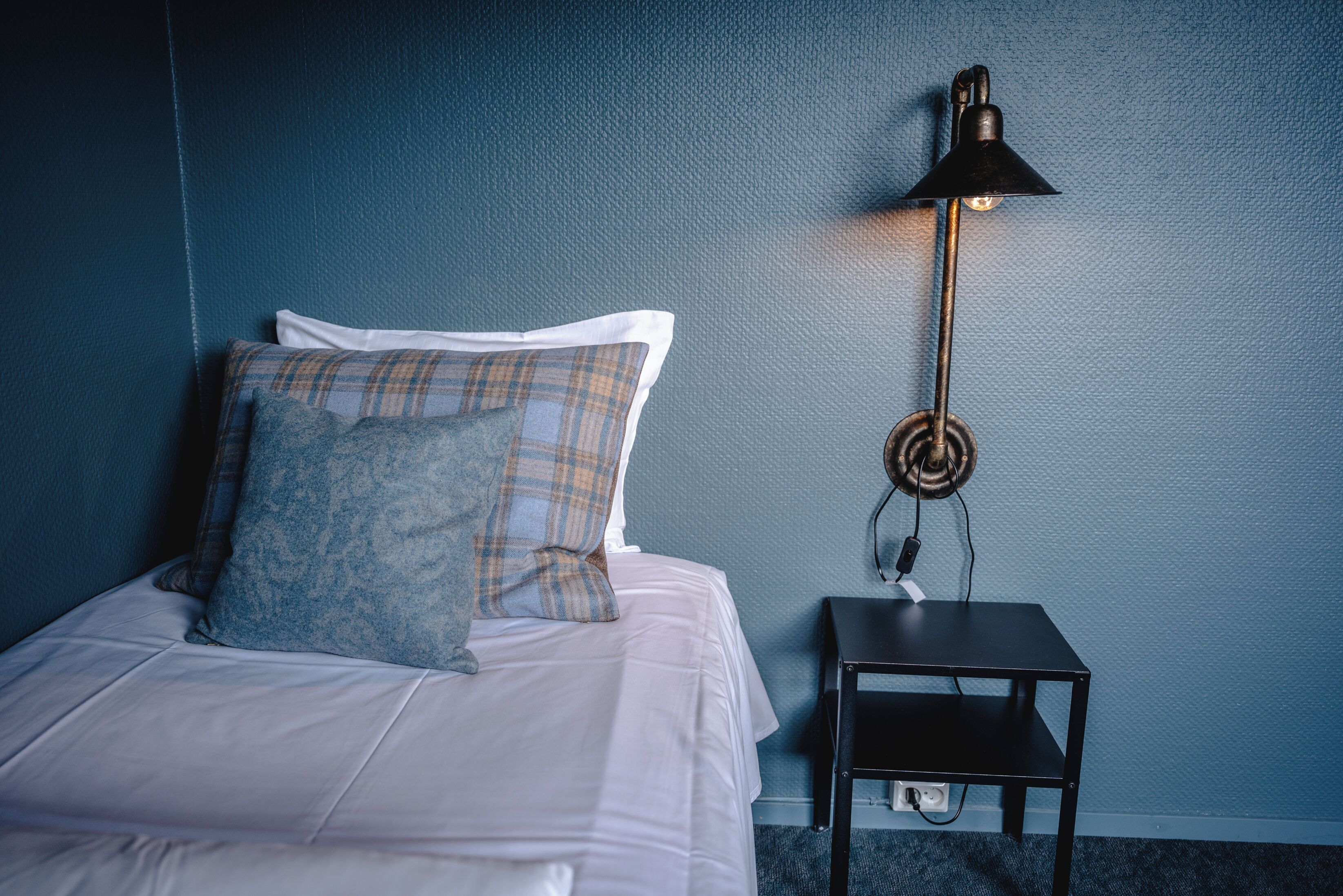 A neatly made bed with white linens and plaid pillows, a black nightstand with a lamp, and a blue textured wall.