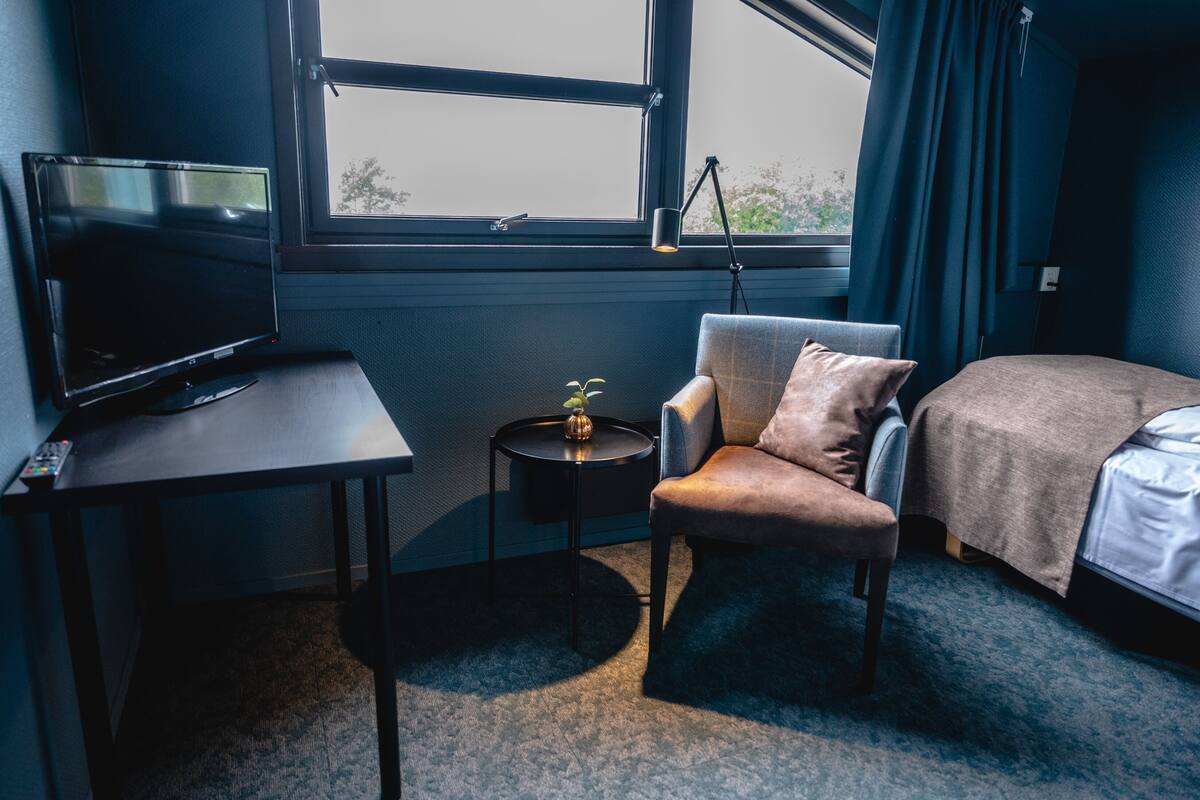 A hotel room with a bed, a chair, a small table with a lamp, and a TV.