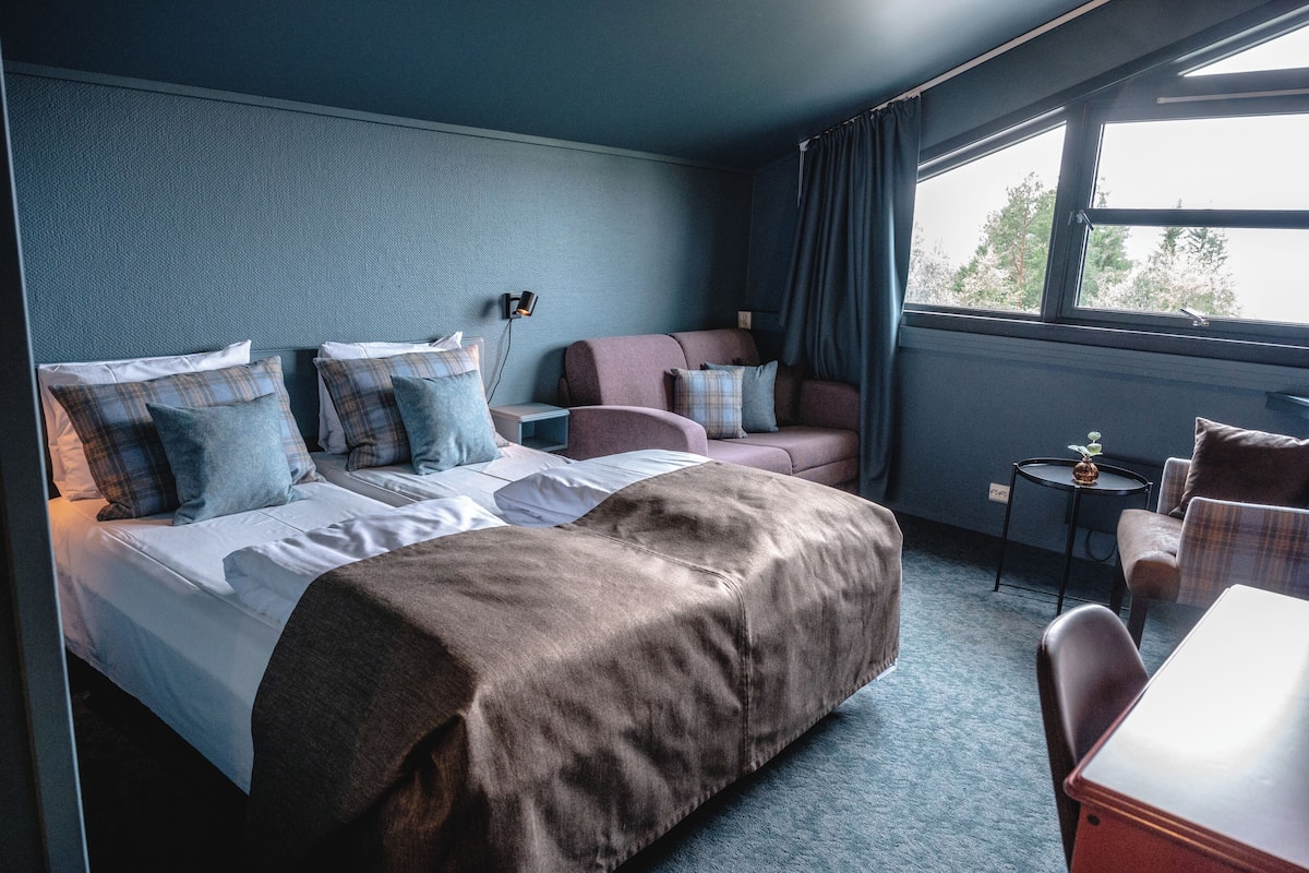 A hotel room with a large bed, a sofa, a small table, and a window with curtains.