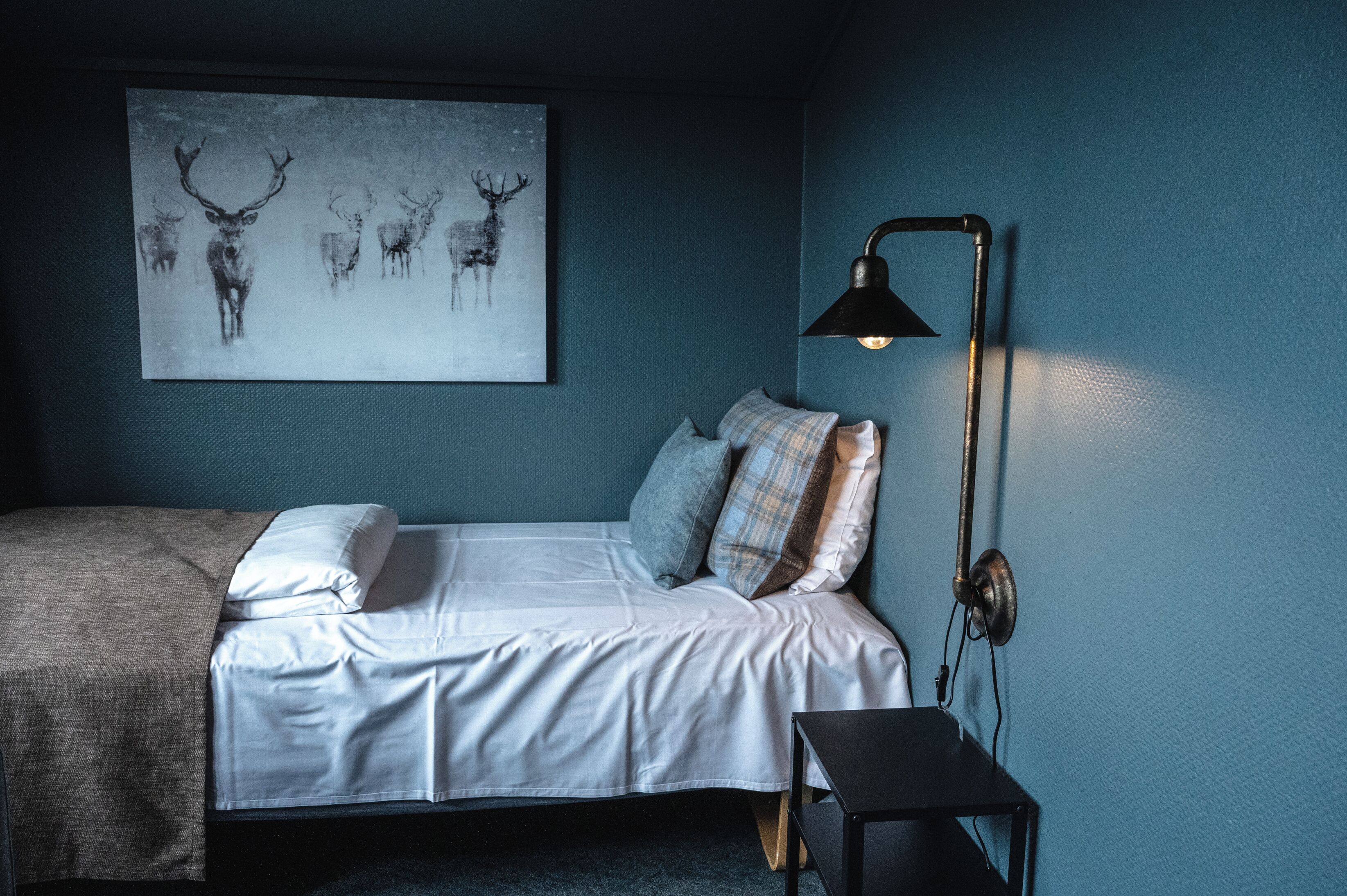 Un dormitorio con cama, almohadas, una mesita de noche, una lámpara de pared y una obra de arte con motivos de ciervo.