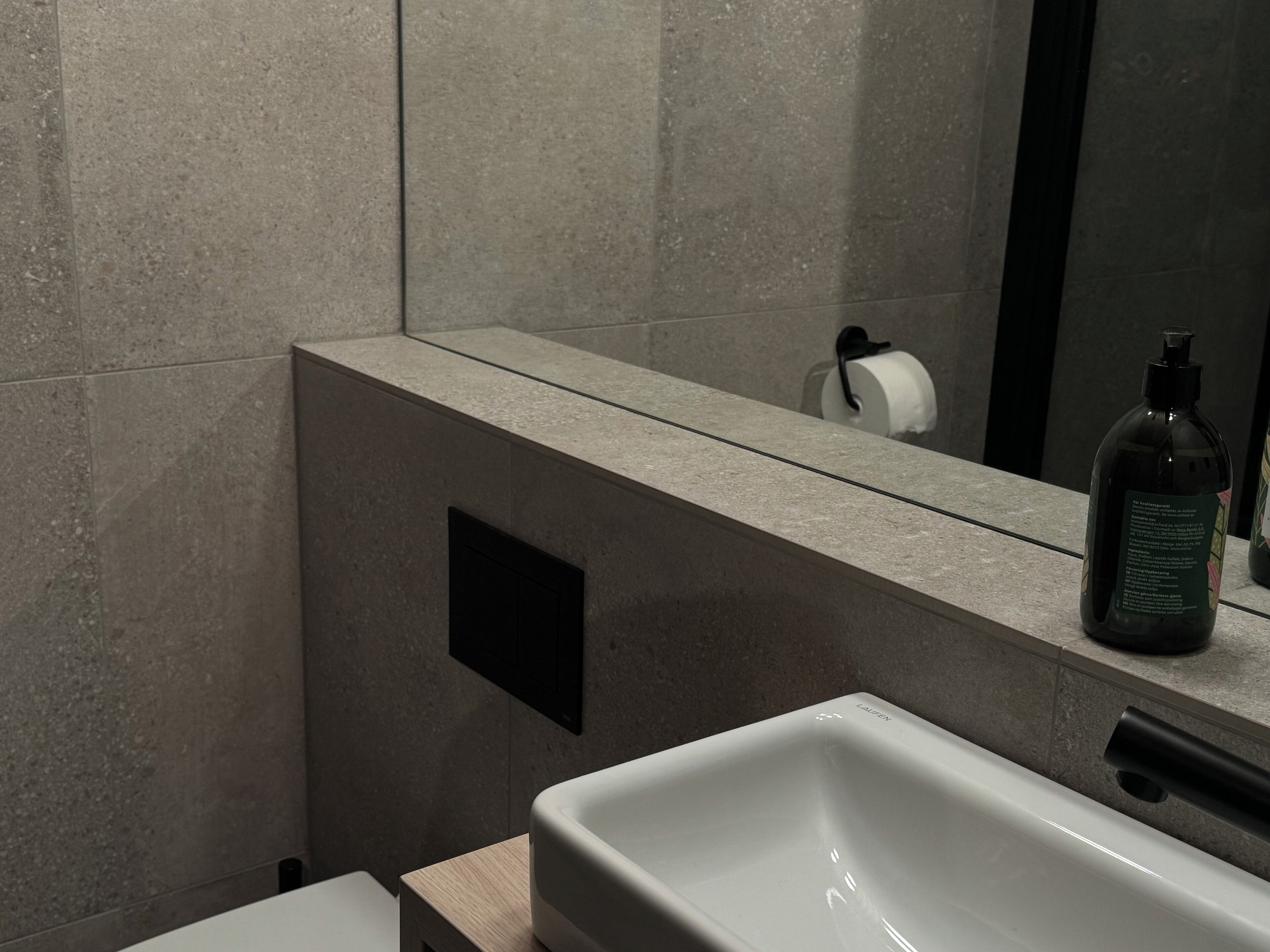 Ein modernes Badezimmer mit einem großen Spiegel, einem weißen Waschbecken und einer Shampooflasche auf der Ablagefläche.