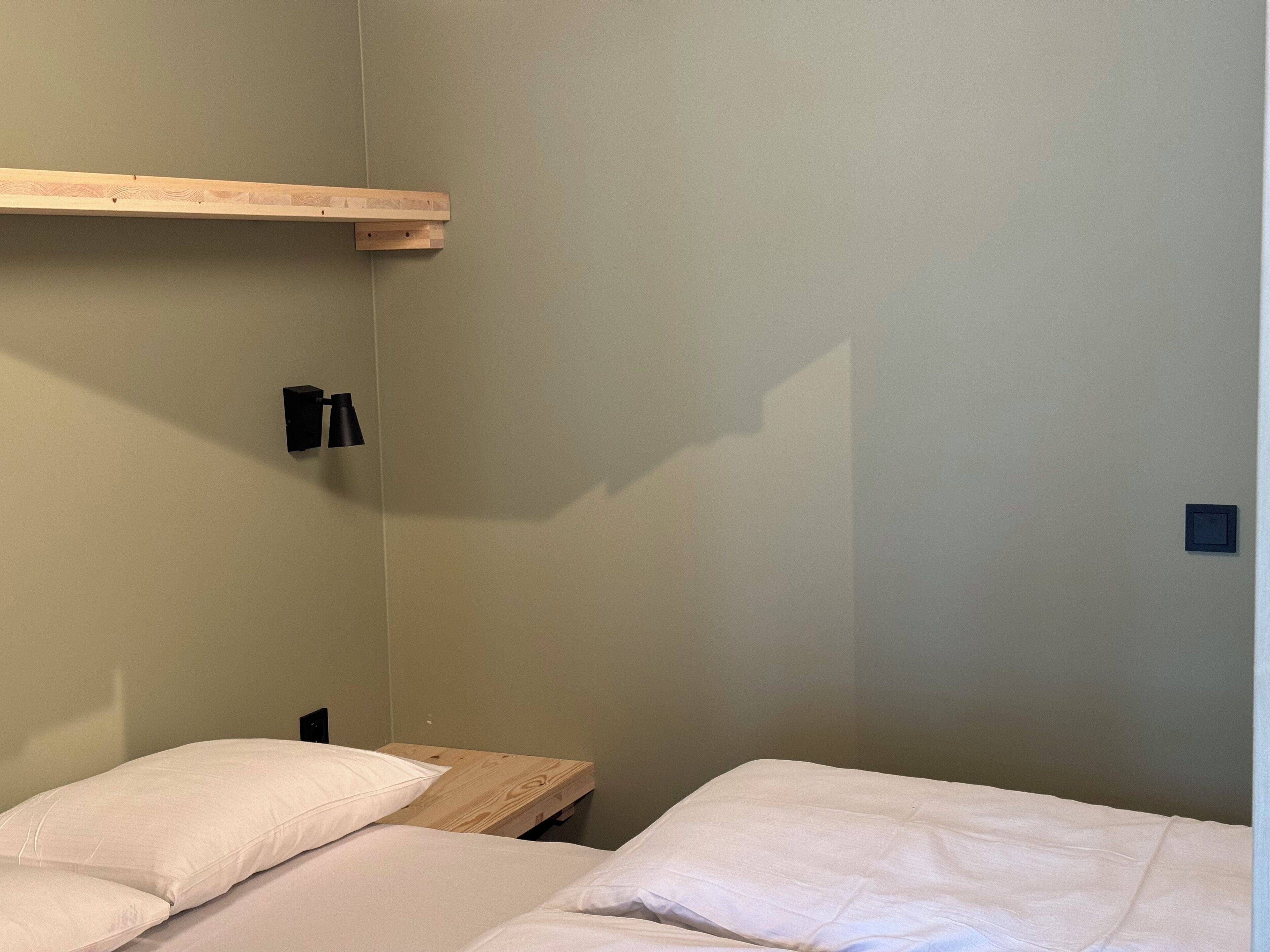 Ein kleiner, schlichter Raum mit einem Bett, einem Holzbodenregal und einer schwarzen Wandleuchte.