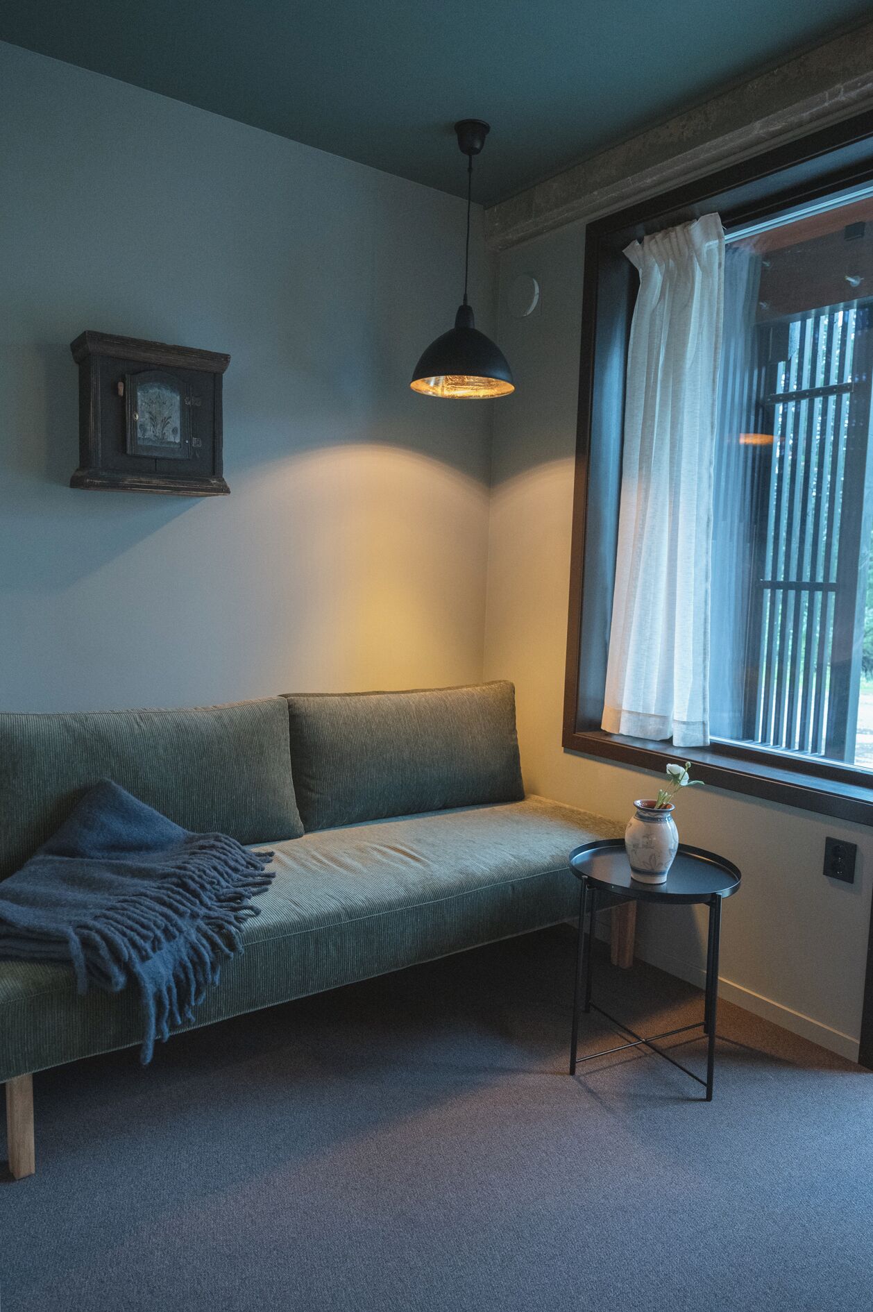 A room with a green sofa, a small table with a vase, a hanging light, and a window with curtains.