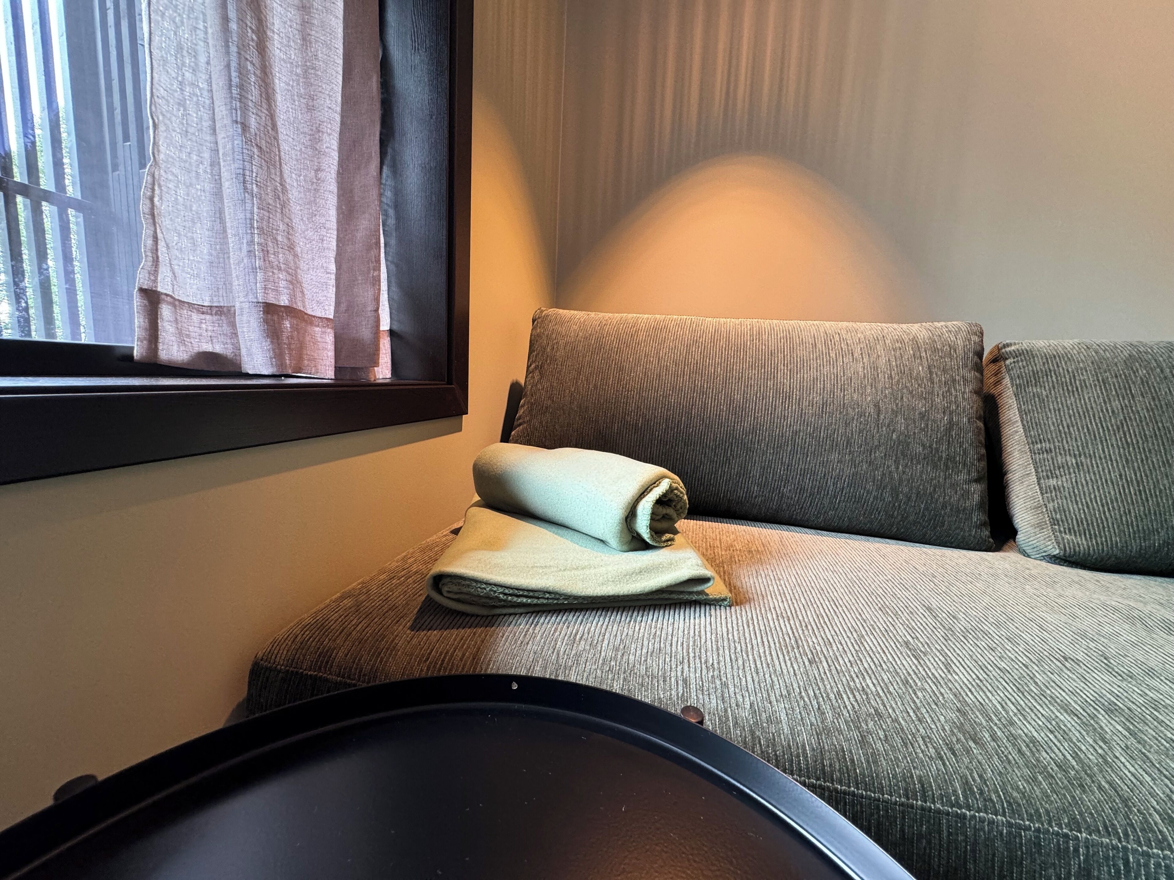 A room with a grey sofa, a window with curtains, and a small table with a folded towel.