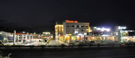 A Visthara Inn building with a well-lit facade, a gas station in front, and a motorcycle parked on the roadside.