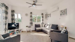 Ein modernes Wohnzimmer mit einem Flachbildfernseher, grauen Sofas, einem kleinen weißen Tisch und einem Deckenventilator.
