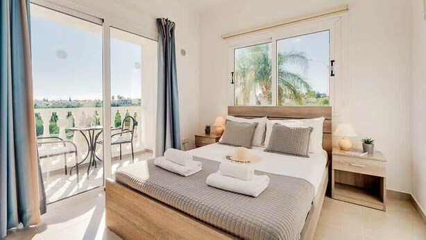 Ein modernes Schlafzimmer mit einem großen Bett, einem Nachttisch, einem Fenster mit Blick und einem Balkon mit Gartenmöbeln.