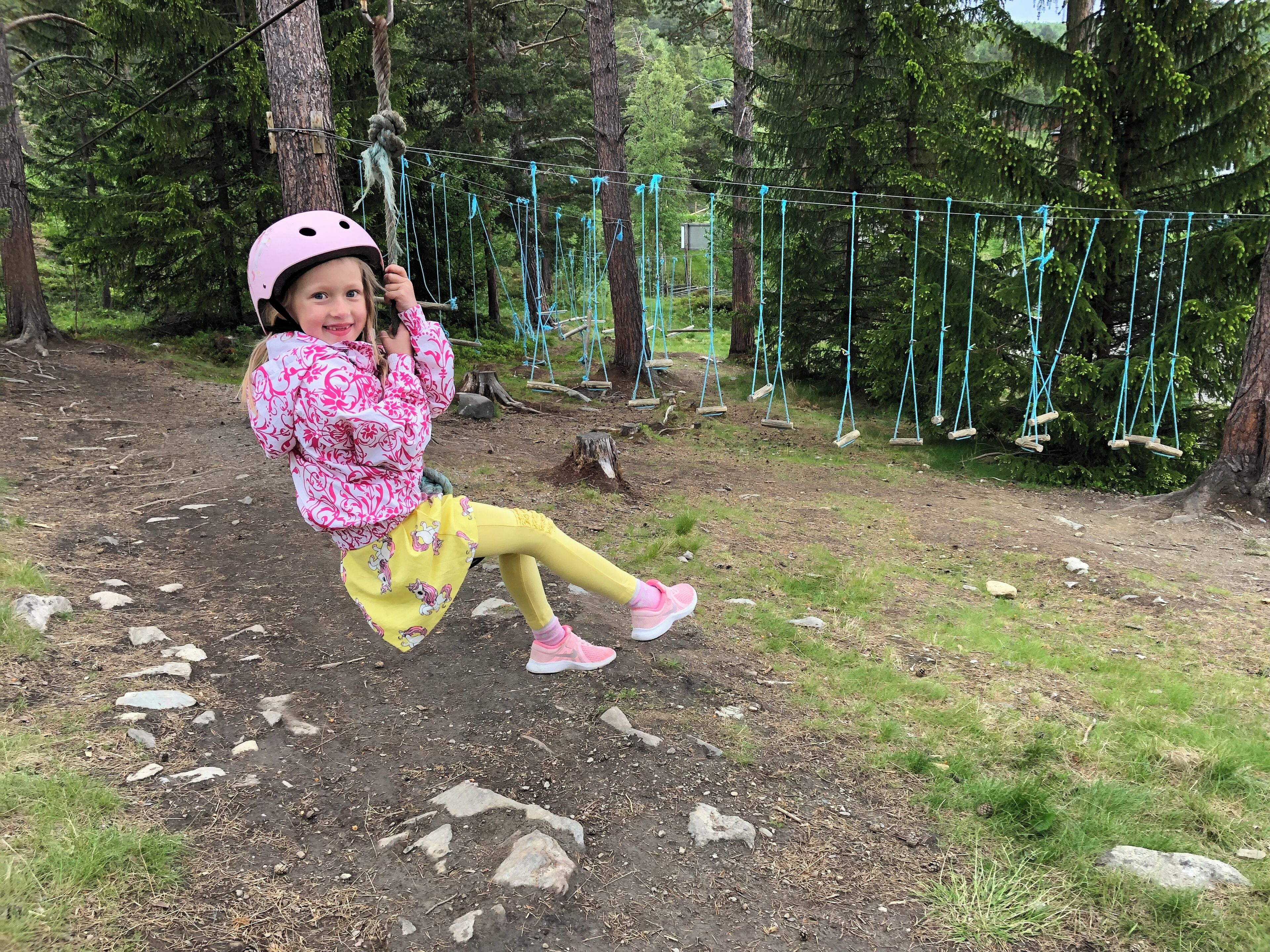 A child on a rope bridge in a forested area with swings and trees in the background.