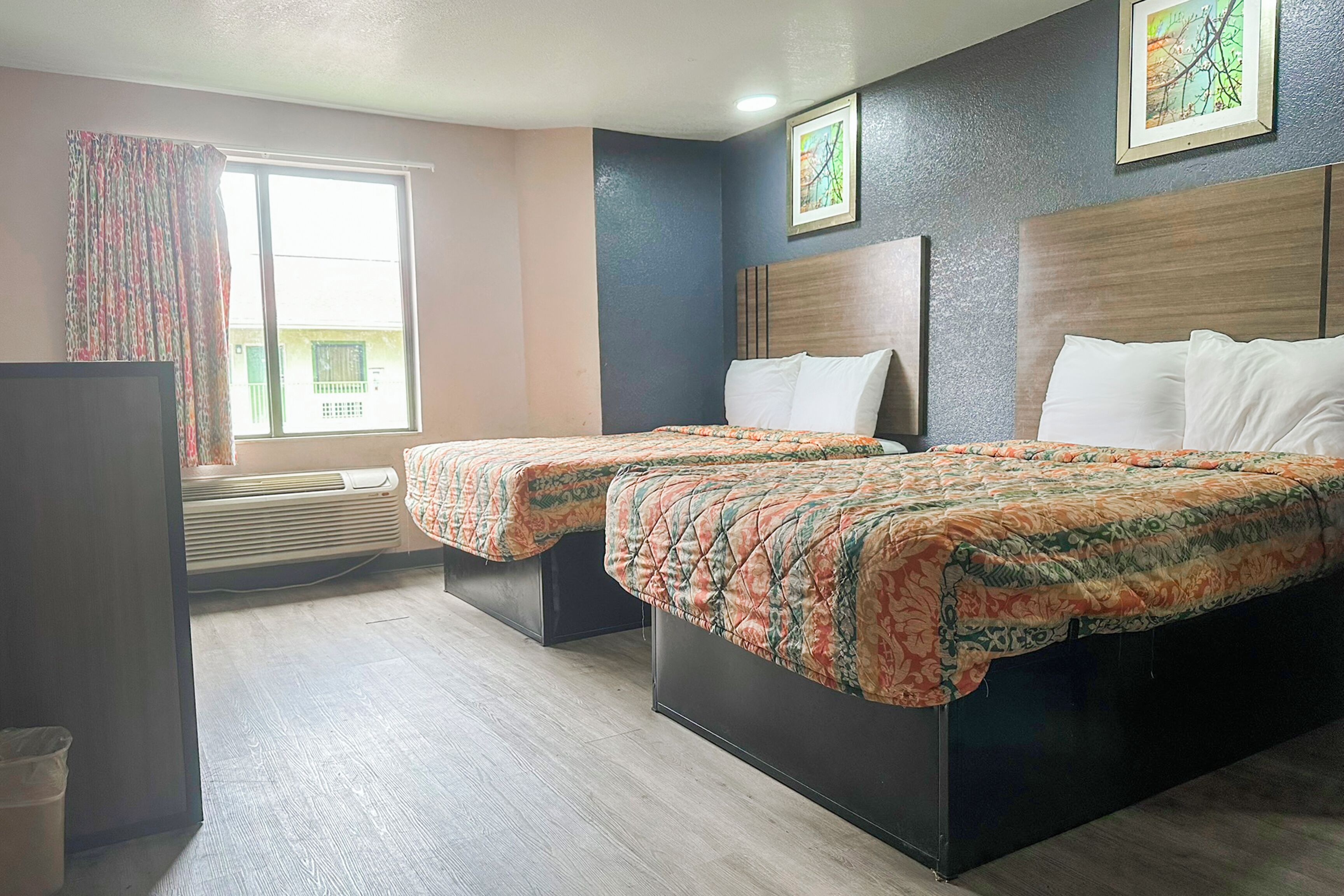 A hotel room with two beds, a large window with curtains, an air conditioning unit, and framed artwork on the wall.