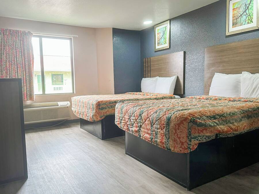 Quarto de hotel com duas camas, uma janela grande com cortinas, um aparelho de ar condicionado e uma obra de arte emoldurada na parede.