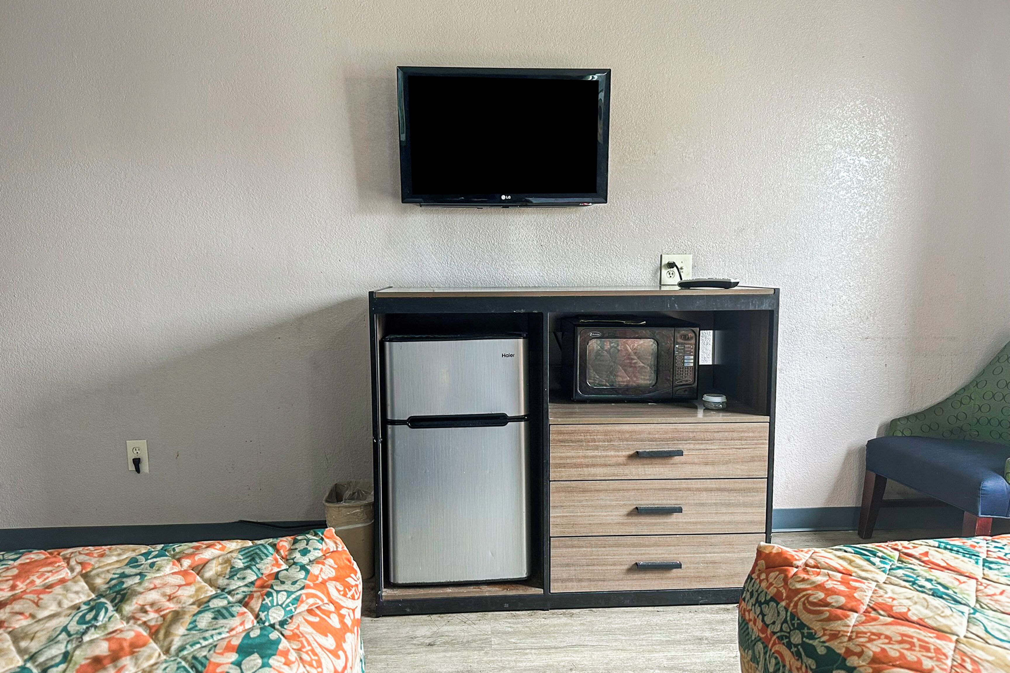 Ein kleines Hotelzimmer mit Küchenzeile, einem Flachbildfernseher, zwei Betten mit gemusterten Bettbezügen und einem grünen Sessel.