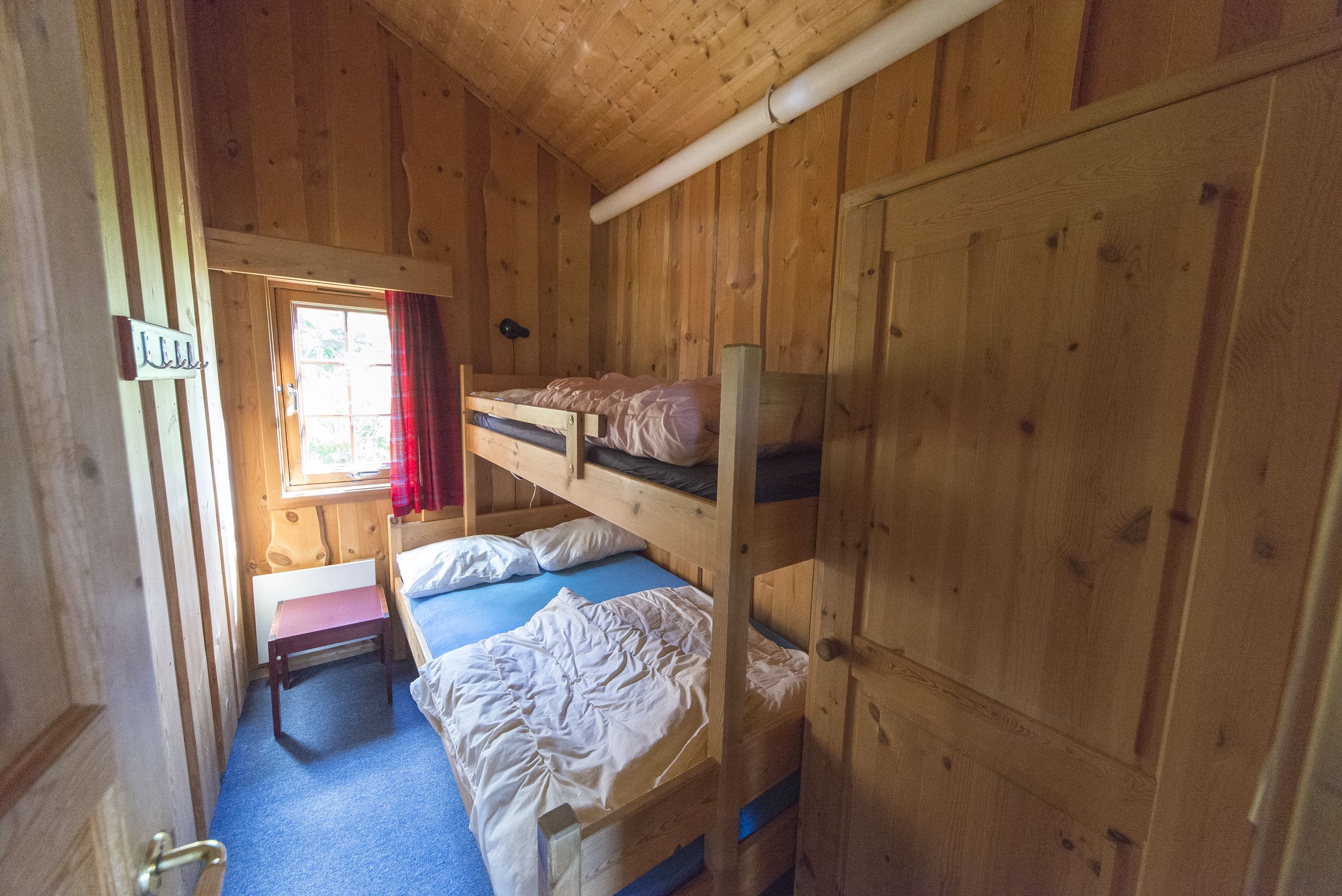 Une chambre dans un chalet en bois, avec des lits superposés, une petite table et une fenêtre avec des rideaux.