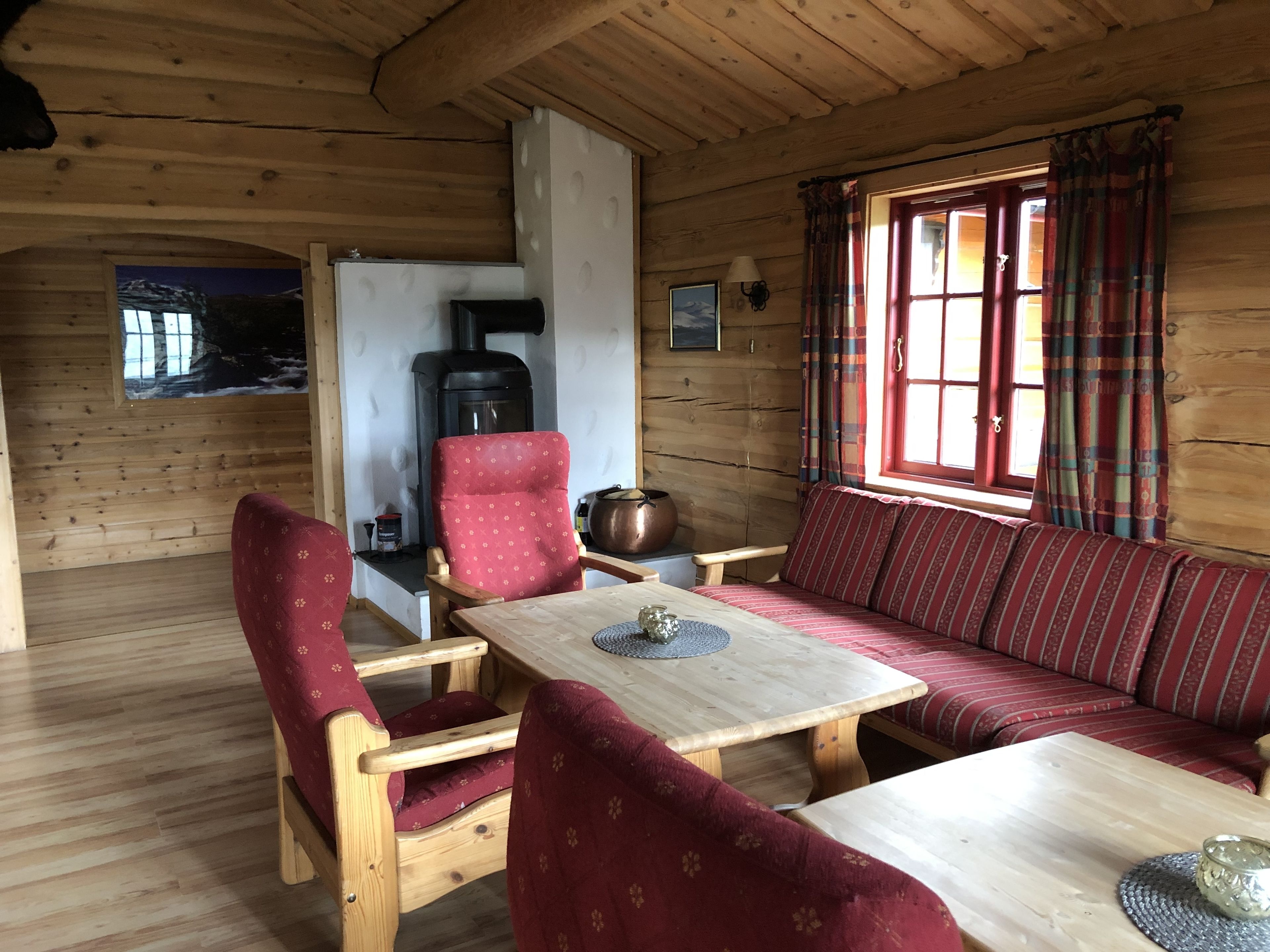A cozy cabin interior with a wooden interior, a sofa, chairs, a table, a fireplace, and a window with curtains.