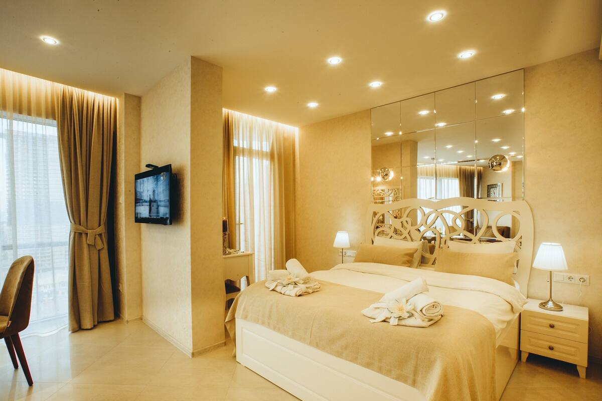 A hotel room with a bed, bedside tables, a TV, a chair, and a large mirror.