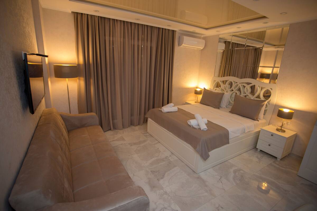 A modern hotel room with a bed, a sofa, a nightstand, a lamp, and a large mirror.