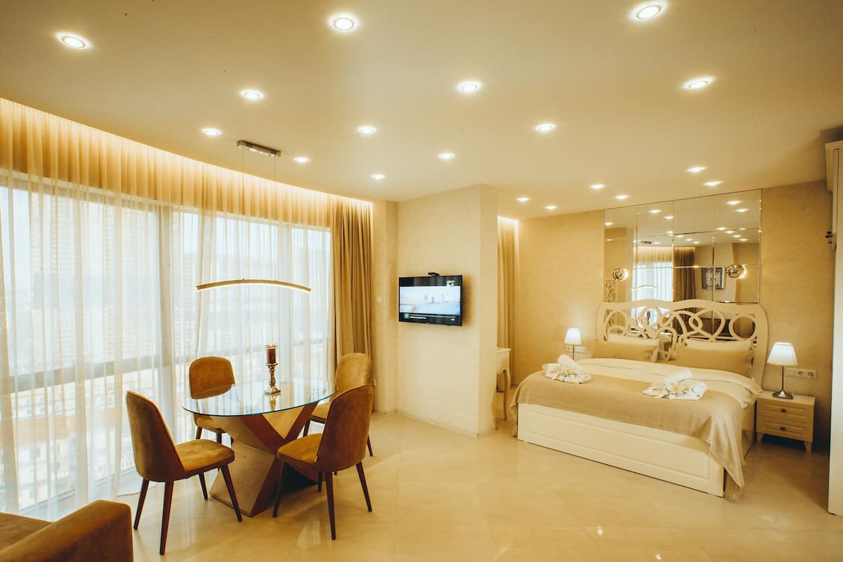 A hotel room with a bed, bedside table, lamp, TV, and a dining area with chairs and a glass table.