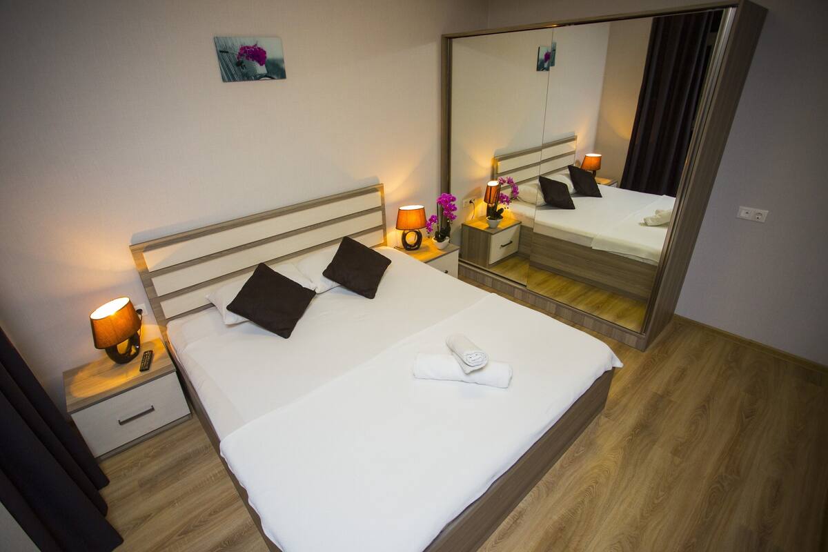 A neatly made bed with a white comforter, two dark pillows, a folded white towel, a bedside lamp, and a nightstand with a remote control.