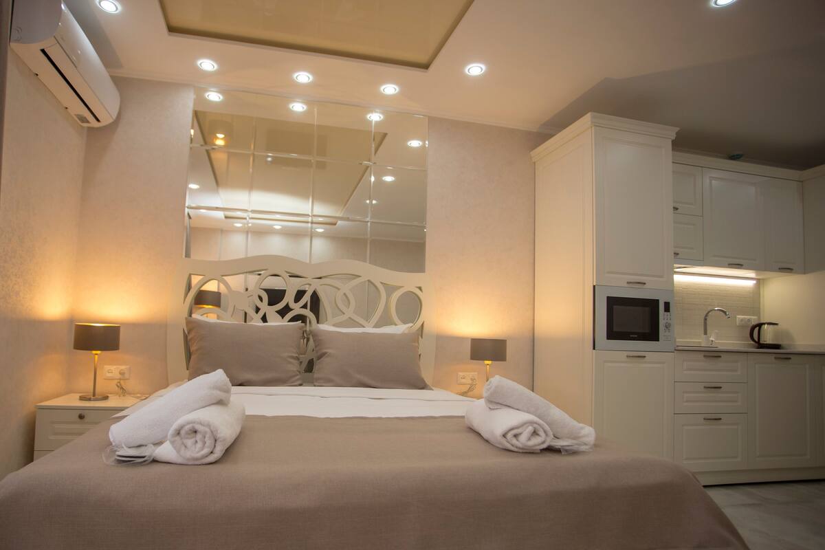 A hotel room with a bed, bedside lamps, a mirror, a kitchenette, and an air conditioning unit.