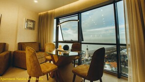Un salon moderne doté d’une table à manger en verre, de sièges confortables et d’une grande fenêtre offrant une vue sur la mer et le paysage urbain.