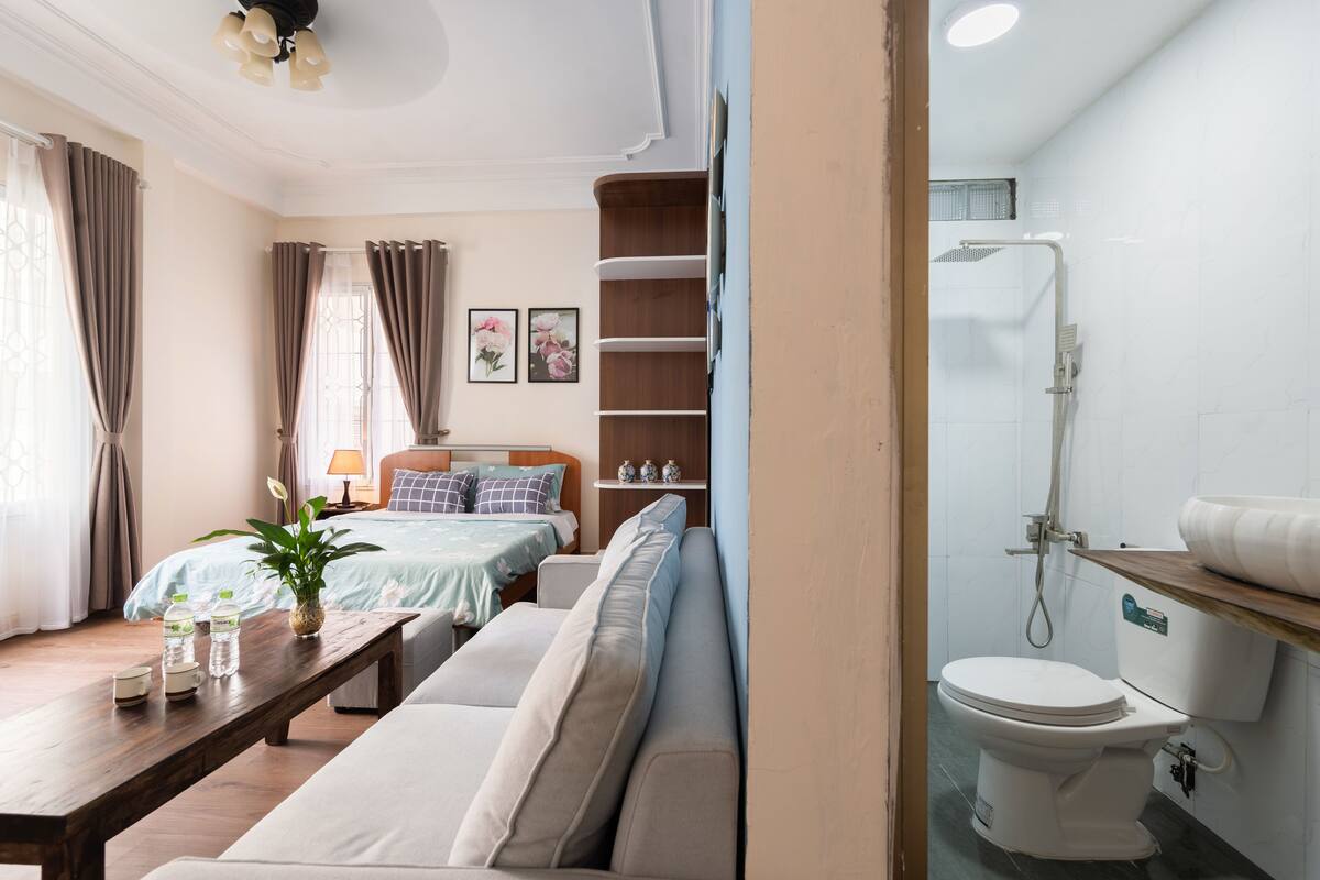 A hotel room with a bed, a sofa, a wooden table with bottles, a potted plant, and a bathroom with a toilet and shower.