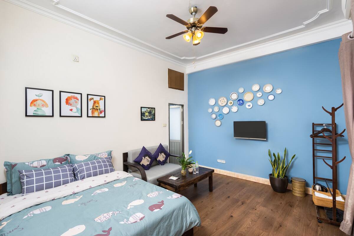 A room with a bed, a sofa, a coffee table, a ceiling fan, a wall-mounted TV, a plant, and a coat rack.