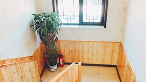 Canto de um quarto com um banco de madeira, uma planta em vaso e uma janela com grades de metal.