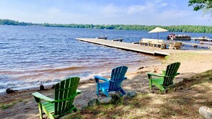 En vacker utsikt över sjön med en brygga som sträcker sig ut i vattnet, där flera båtar ligger förtöjda och en rad gröna Adirondack-stolar står uppradade längs stranden.