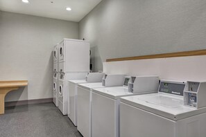 A laundry facility with multiple washing machines and dryers, a wooden bench, and a wall-mounted control panel. - Home2 Suites by Hilton Pecos (Pecos)