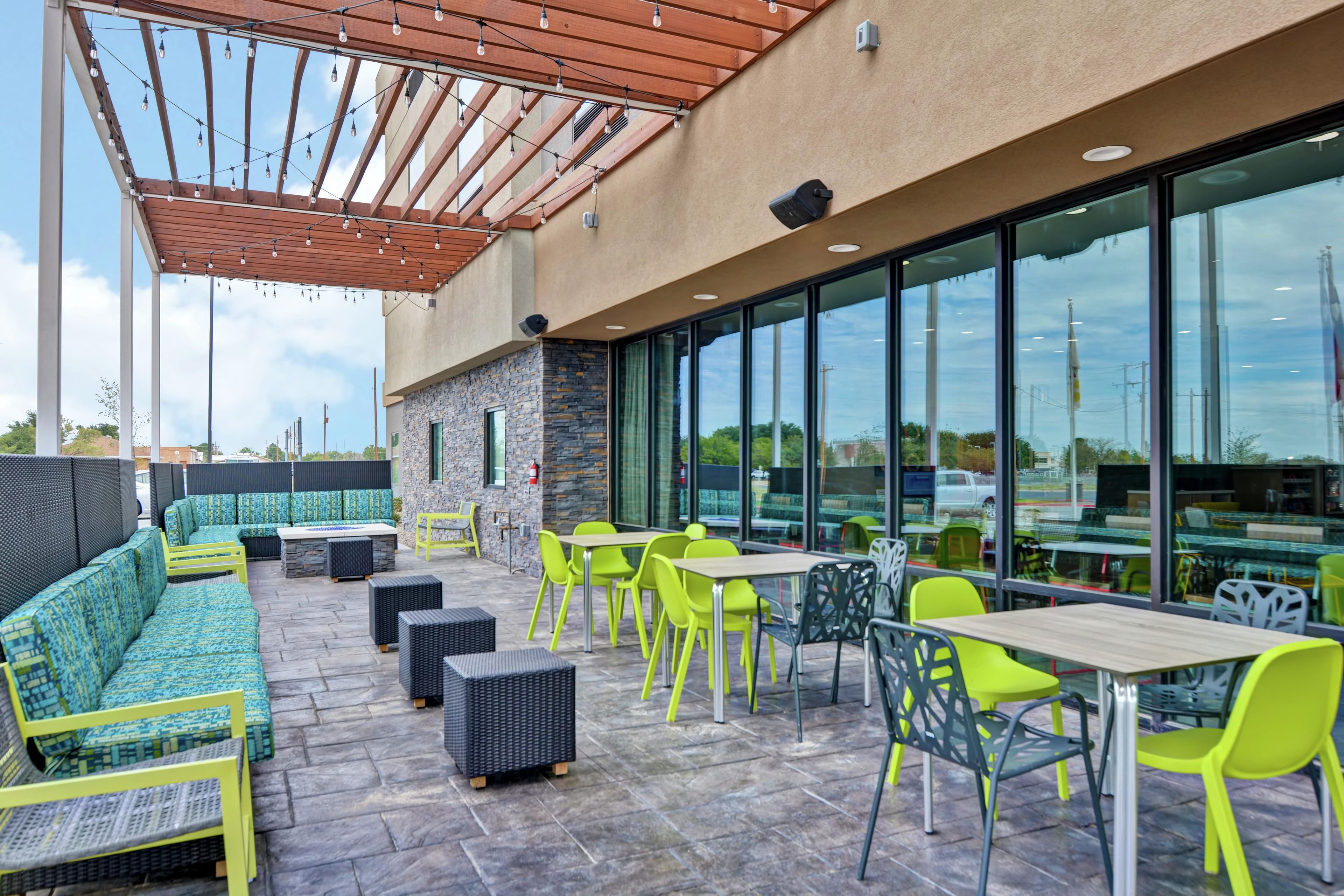 Un patio exterior con disposición de asientos, una pérgola con luces colgantes y vistas a través de grandes ventanales.