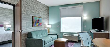 A hotel room with a bed, a sofa, a small table, a chair, a TV, and a window with blinds.