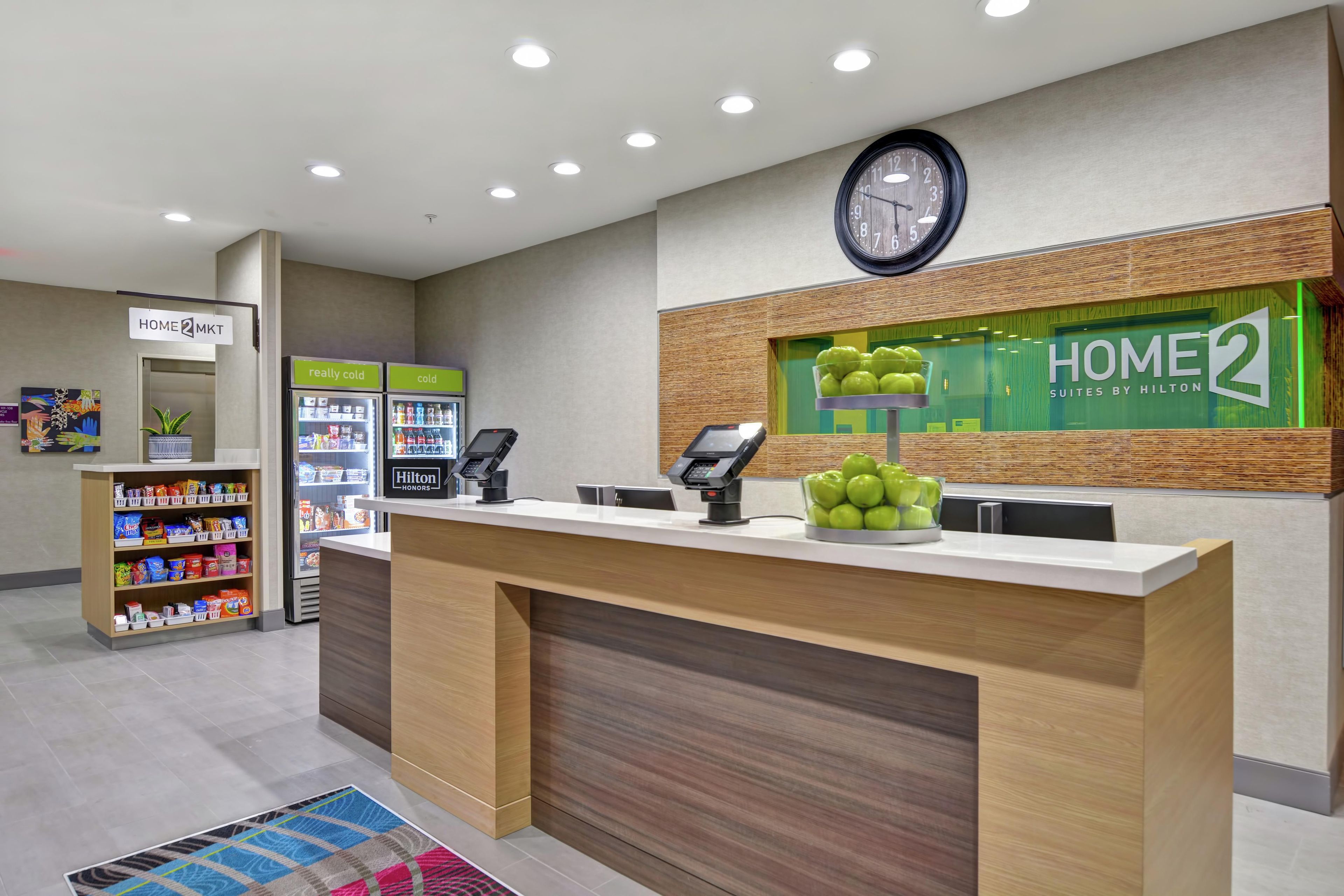 Zona de recepción de un hotel con mostrador, máquinas expendedoras, un reloj y un letrero que dice 'Home 2 Suites by Hilton'.