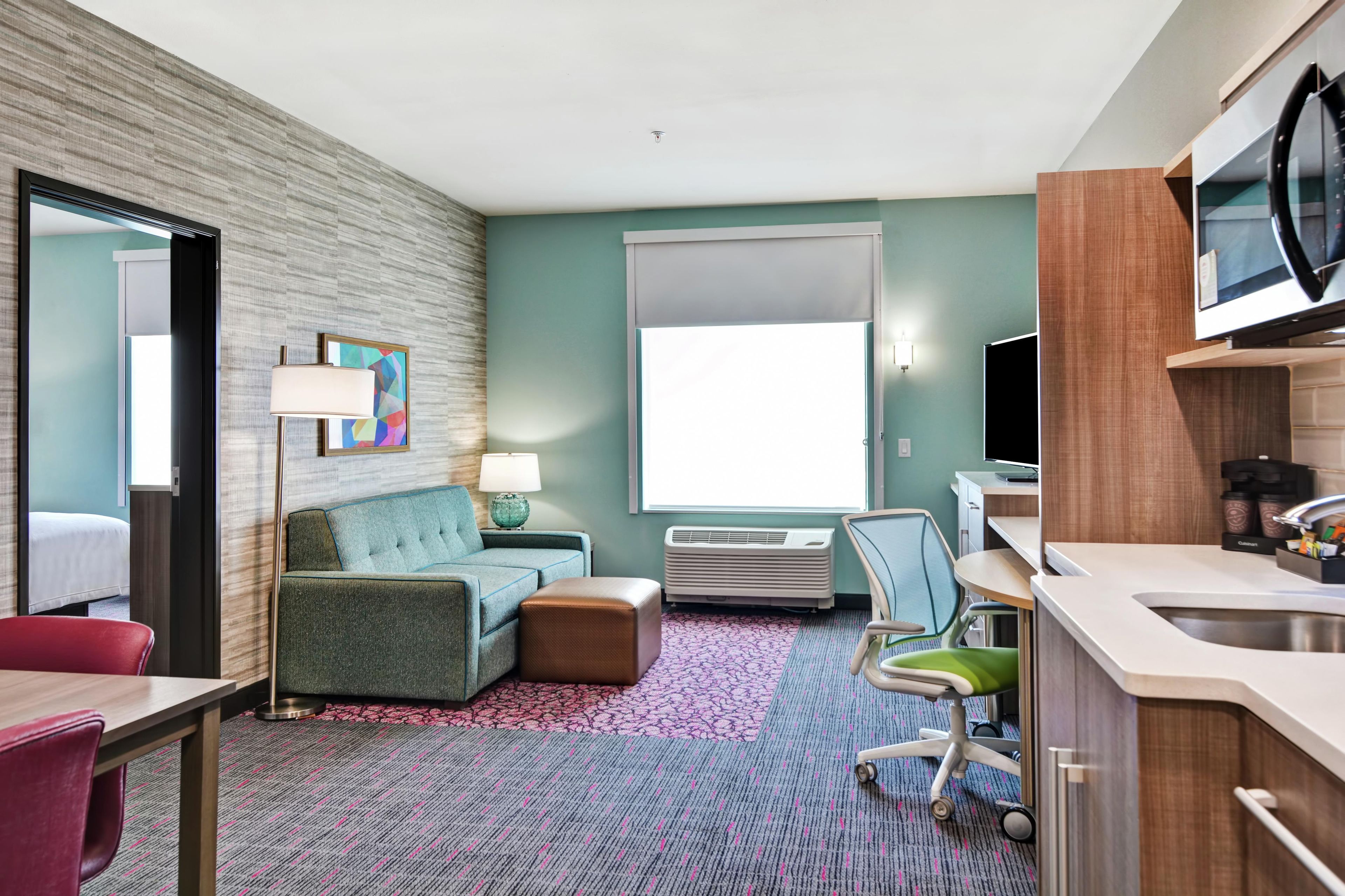 A hotel room with a sofa, a small table, a chair, a desk, a television, a window, and a kitchenette.